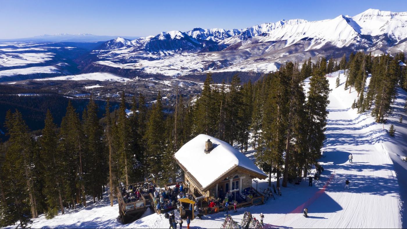 The Alpino Vino chalet on the See Forever run at Telluride Ski Resort, its deck and stone facade among the pines with the San Juan peaks behind at 11,966 feet