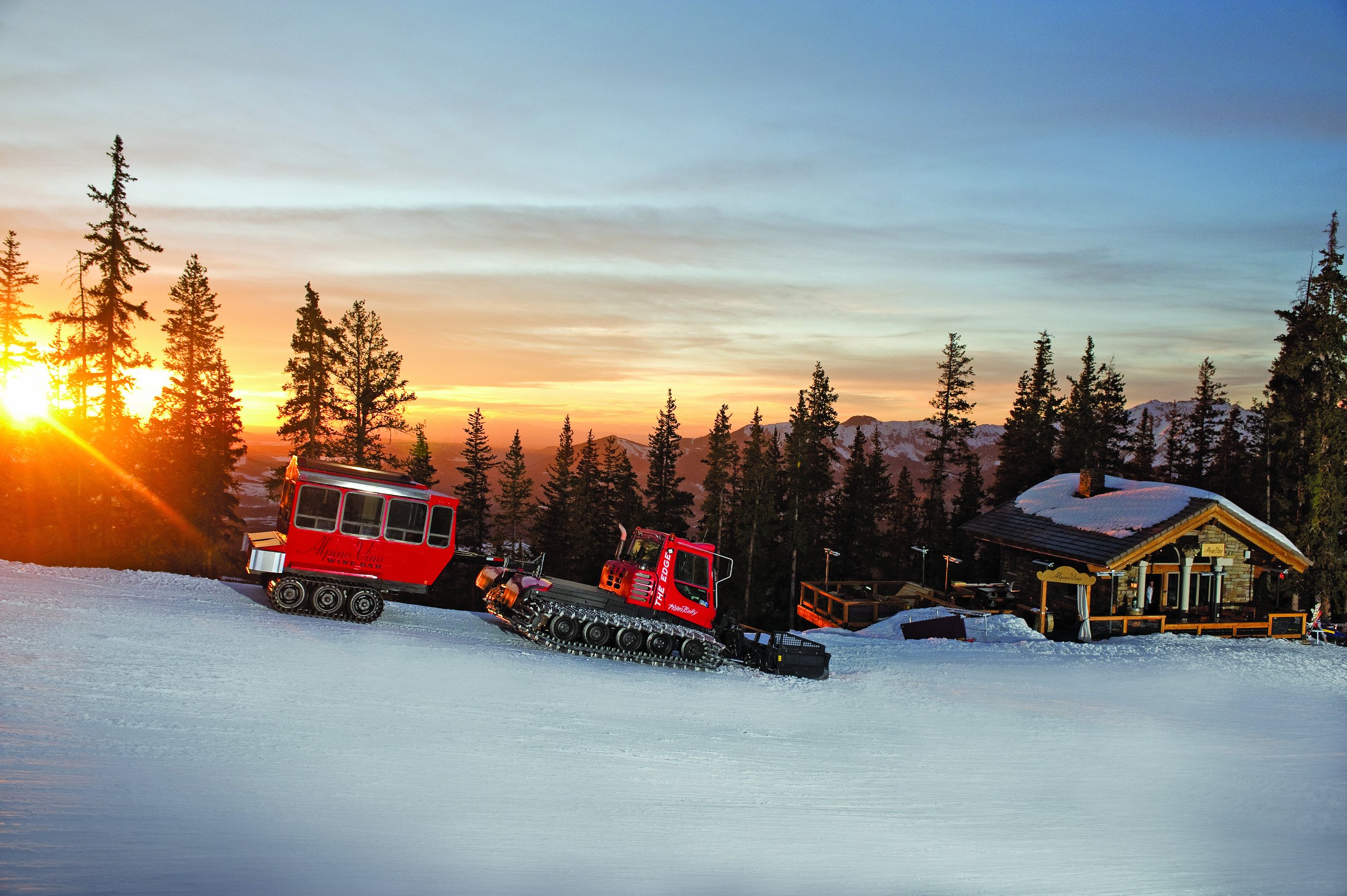 The Alpino Vino Wine Bar snowcoach and a groomer parked on the snow outside the lit stone hutte at sunset, the San Juan peaks beyond, 11,966 feet above Telluride