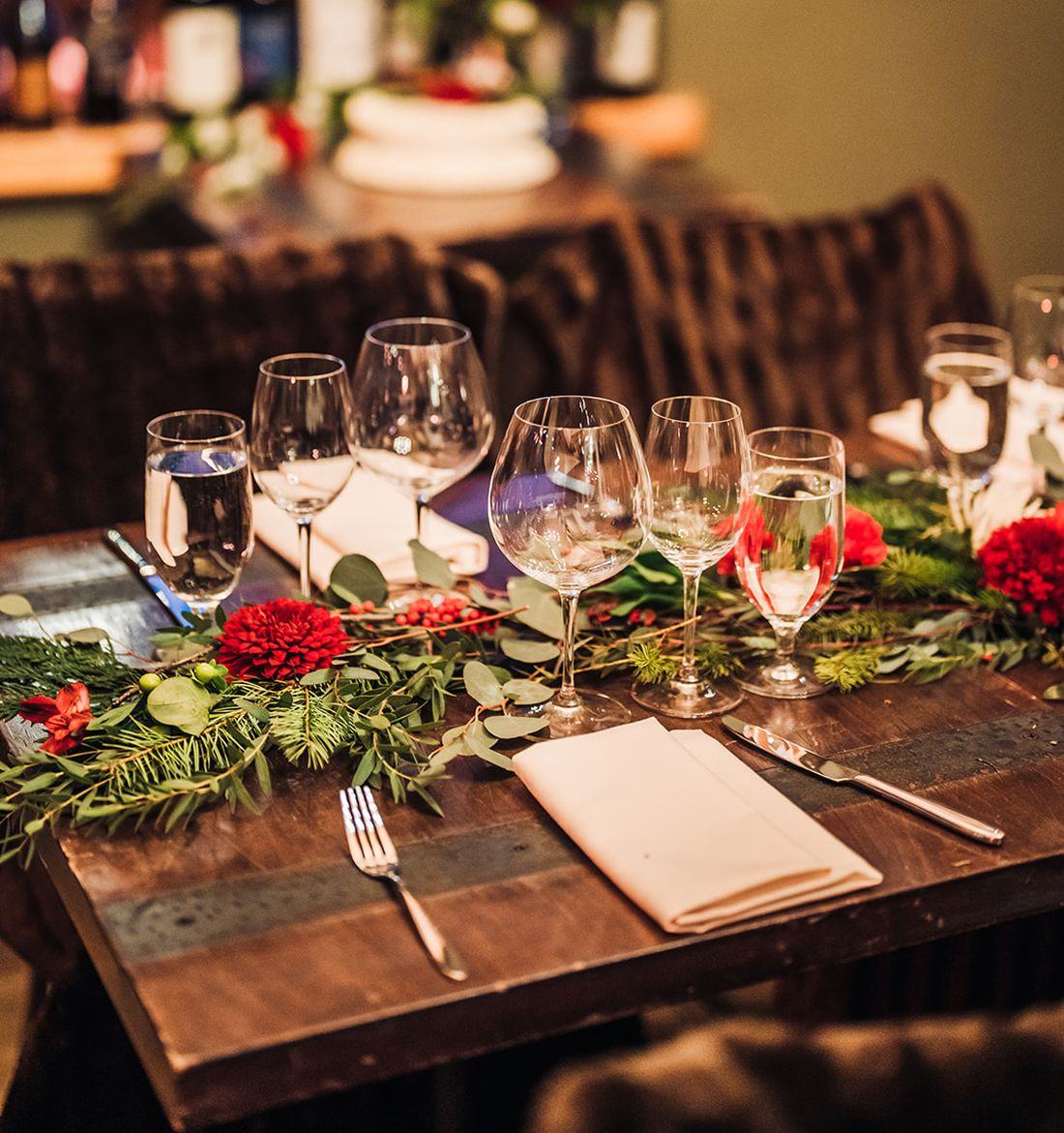 An evening place setting at Alpino Vino: wine glasses, a winter garland of greenery and red blooms, candlelight and the wine shelf behind