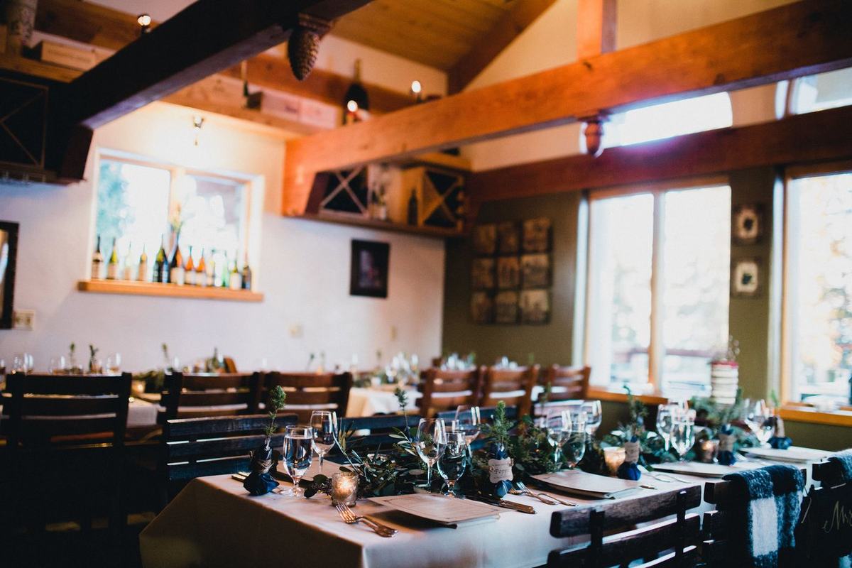 The Alpino Vino dining room set for a private event: one long table with a greenery runner, candles, place cards and blue napkins, beneath hand-hewn beams and the wine racks