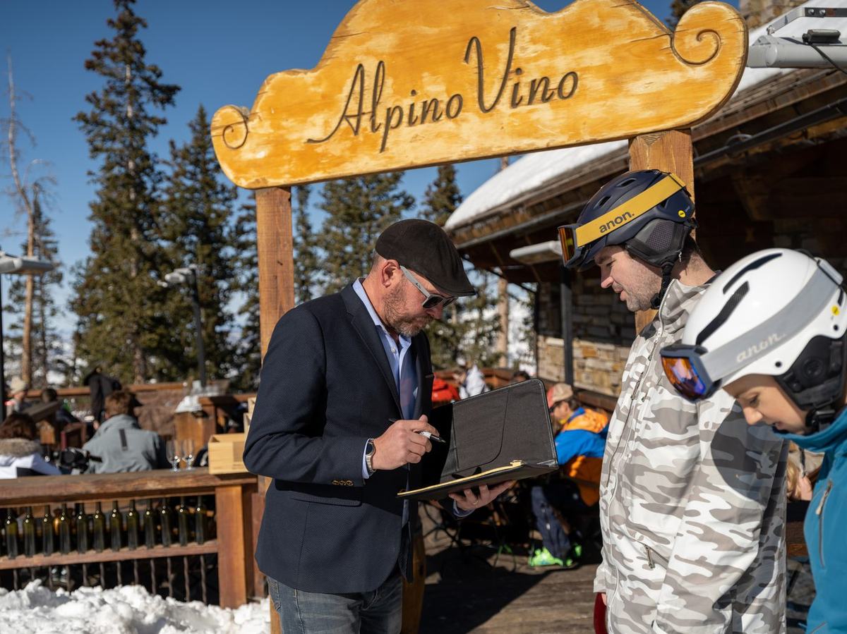 Skiers in helmets checking in with a host under the carved Alpino Vino sign on the sunlit deck at 11,966 feet, Telluride Ski Resort