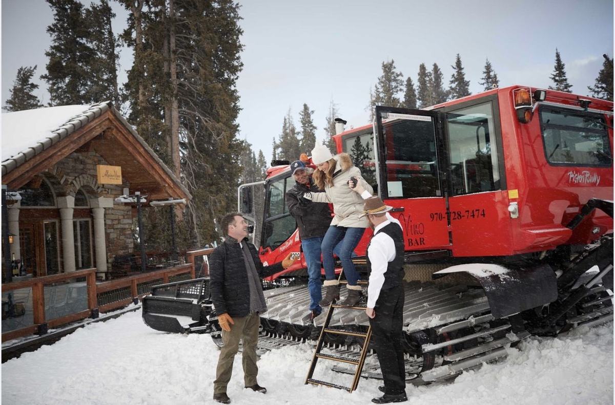 A guest stepping down from the heated Alpino Vino snowcoach onto the snow at the restaurant door, helped by staff, Telluride Ski Resort