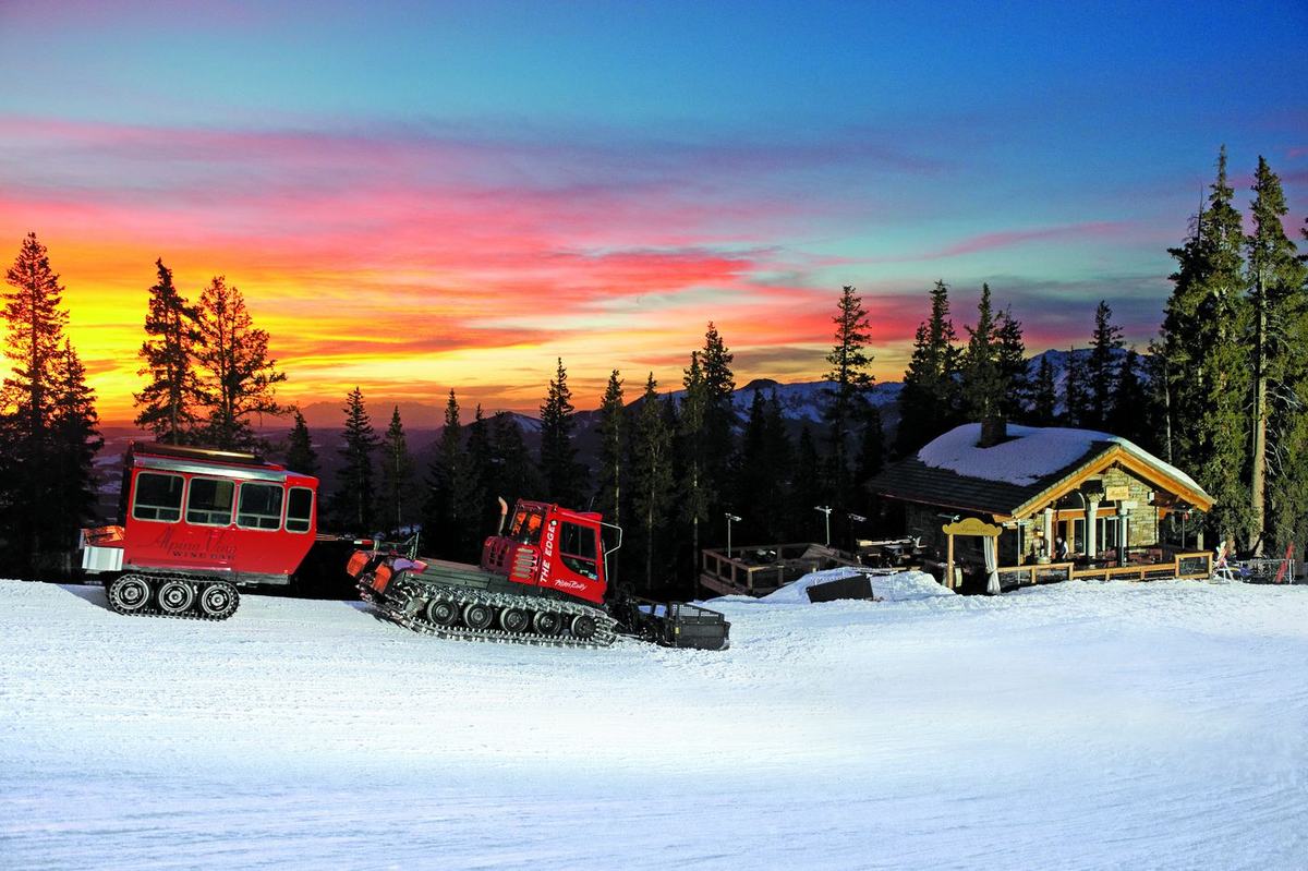 The Alpino Vino snowcoach and its enclosed passenger coach parked outside the lit restaurant at sunset, the sky burning orange over the San Juan peaks at 11,966 feet above Telluride