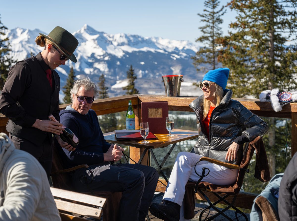 A server presenting a bottle of sparkling rose tableside on the sun deck at Alpino Vino, with the Wilson Range behind at 11,966 feet, Telluride