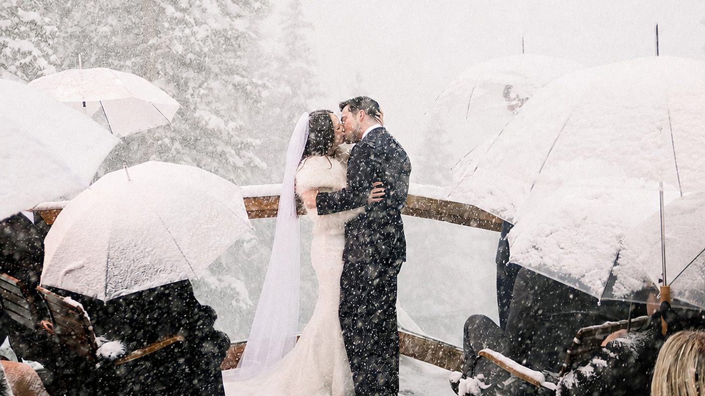 A couple kissing at the end of their ceremony on the Alpino Vino deck in heavy snowfall, guests seated beneath snow-covered umbrellas, snow-laden pines behind, at 11,966 feet above Telluride