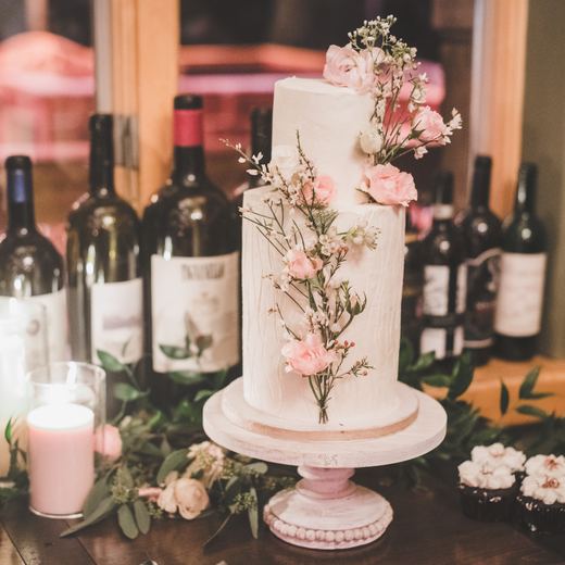 A two-tier wedding cake dressed with pink blooms, set against the wine bottles of the Alpino Vino cellar