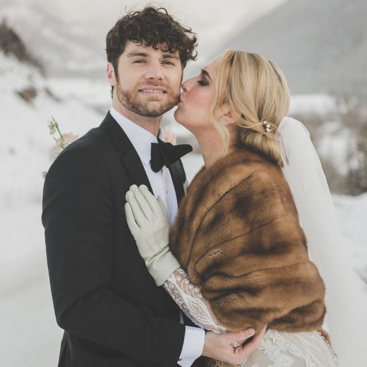 A bride in a fur stole kissing her groom on the cheek in the snow above Telluride