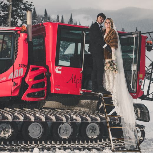 A bride and groom standing in the doorway of the red Alpino Vino snowcoach in falling snow, the restaurant behind them