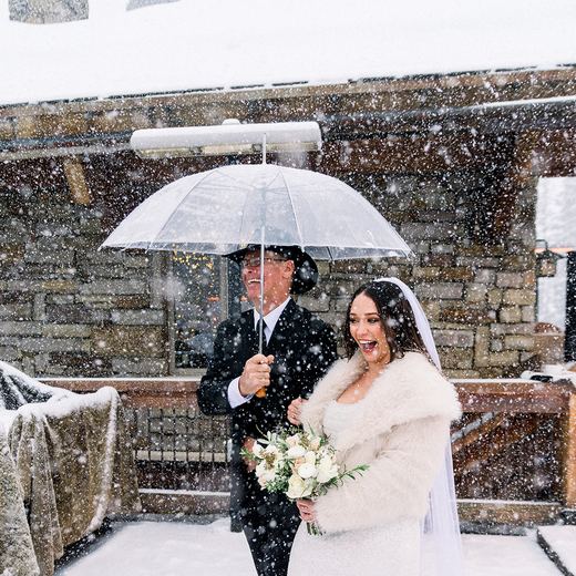 A bride walking in under an umbrella through heavy snowfall at Alpino Vino, escorted past the stone wall of the restaurant
