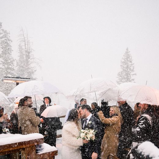 A couple kissing at the end of their ceremony on the Alpino Vino deck in heavy snowfall, guests under umbrellas around them