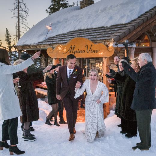 A sparkler send-off beneath the carved Alpino Vino sign at dusk, guests either side of the couple