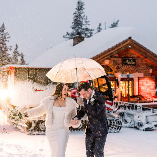 A couple under an umbrella in falling snow beside the snowcoach, sparklers lit and the restaurant glowing behind