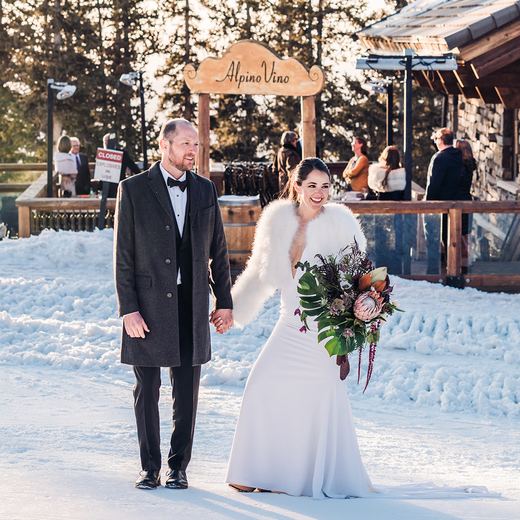 A bride and groom walking through the snow beneath the carved Alpino Vino sign, guests on the deck behind them